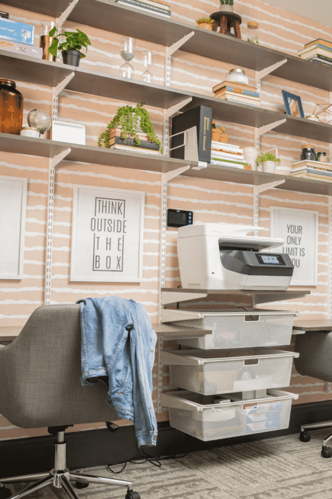 Wall shelving above desks holding up a printer, books, and plastic storage containers.